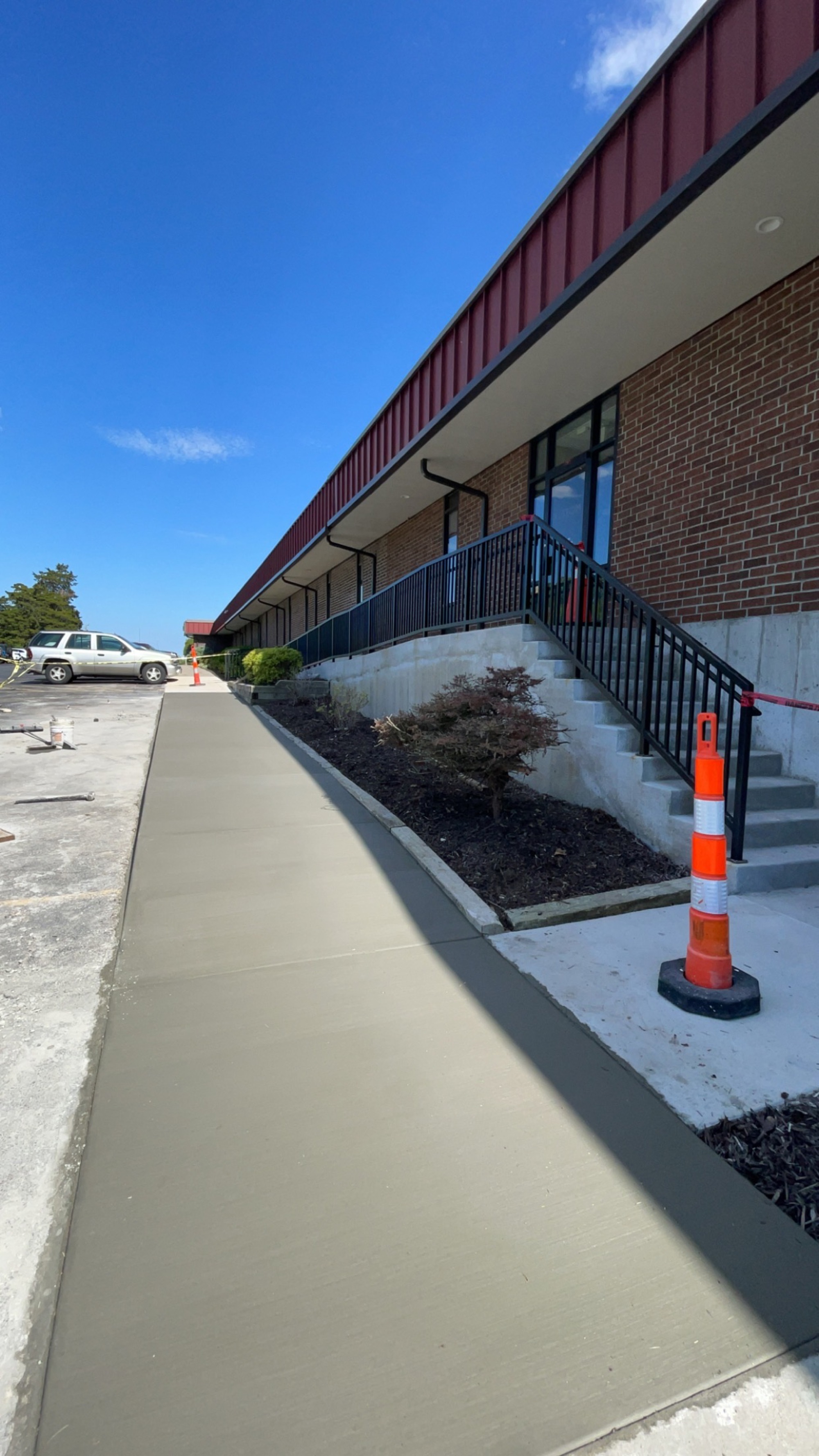 Long finished concrete walkway along a brick commercial building with a red metal roof and covered entrance stairs.