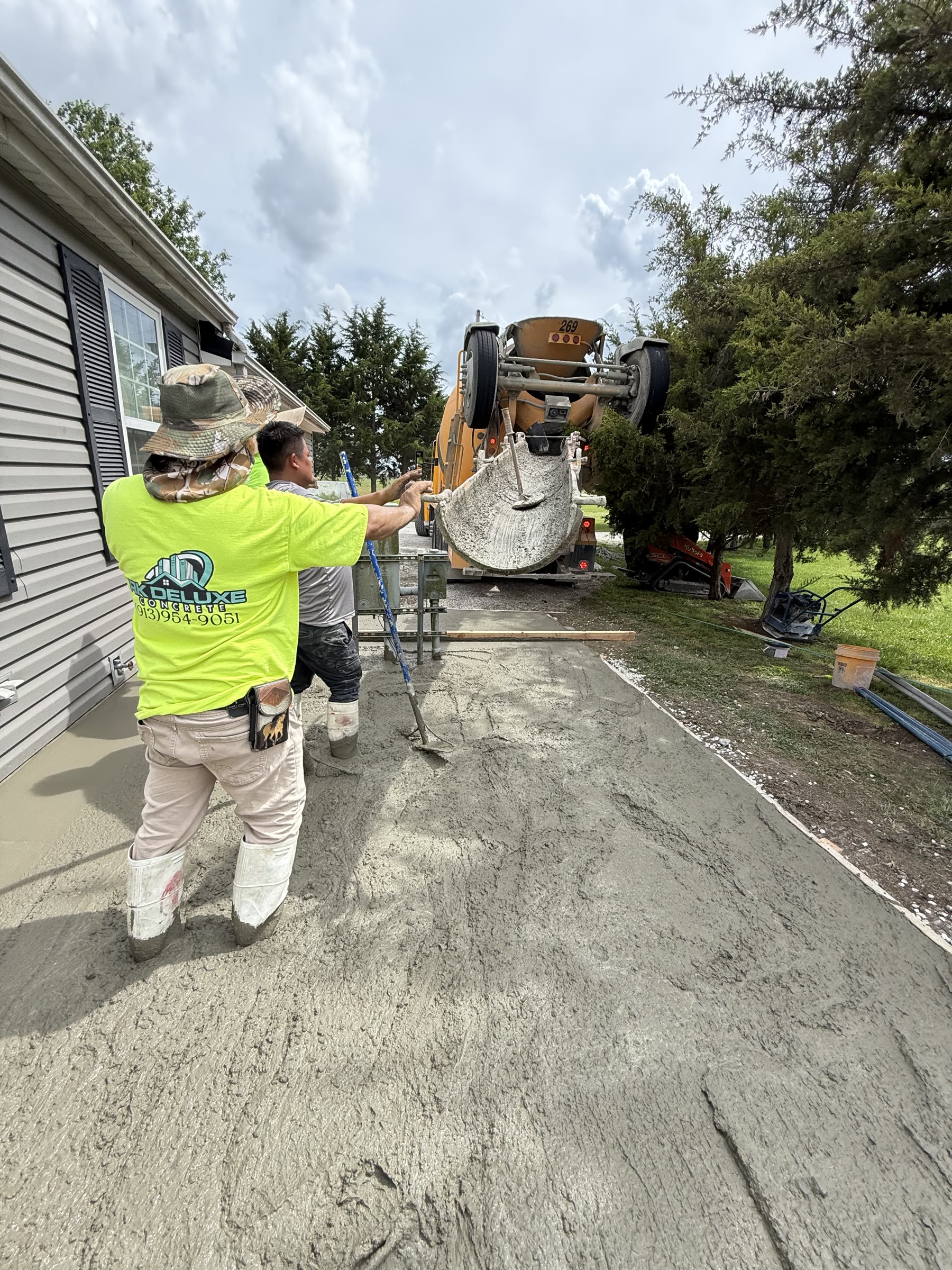 RK Deluxe Concrete crew member in branded work shirt guiding the concrete chute during a pour, mixer truck in background