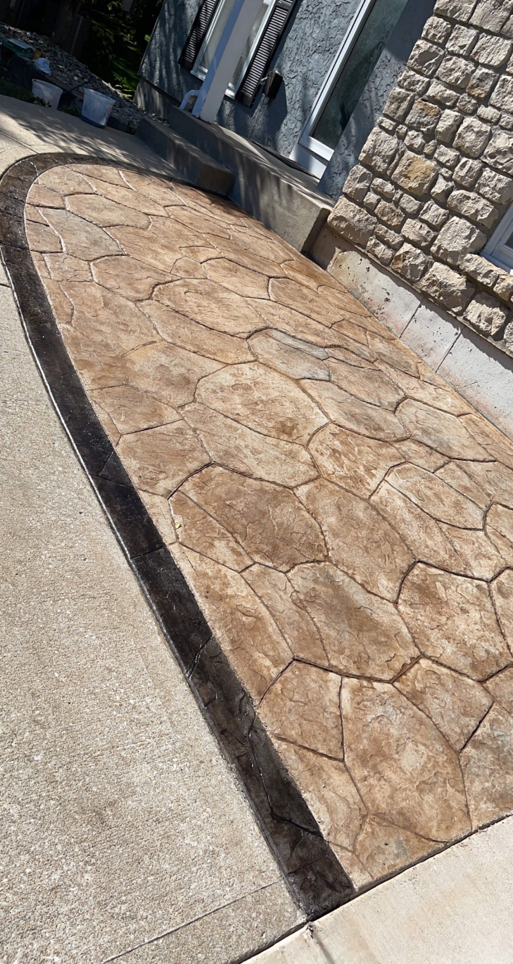 Wide view of an oval stamped-concrete patio in a tan flagstone pattern with a dark stained border, next to a stone house wall.