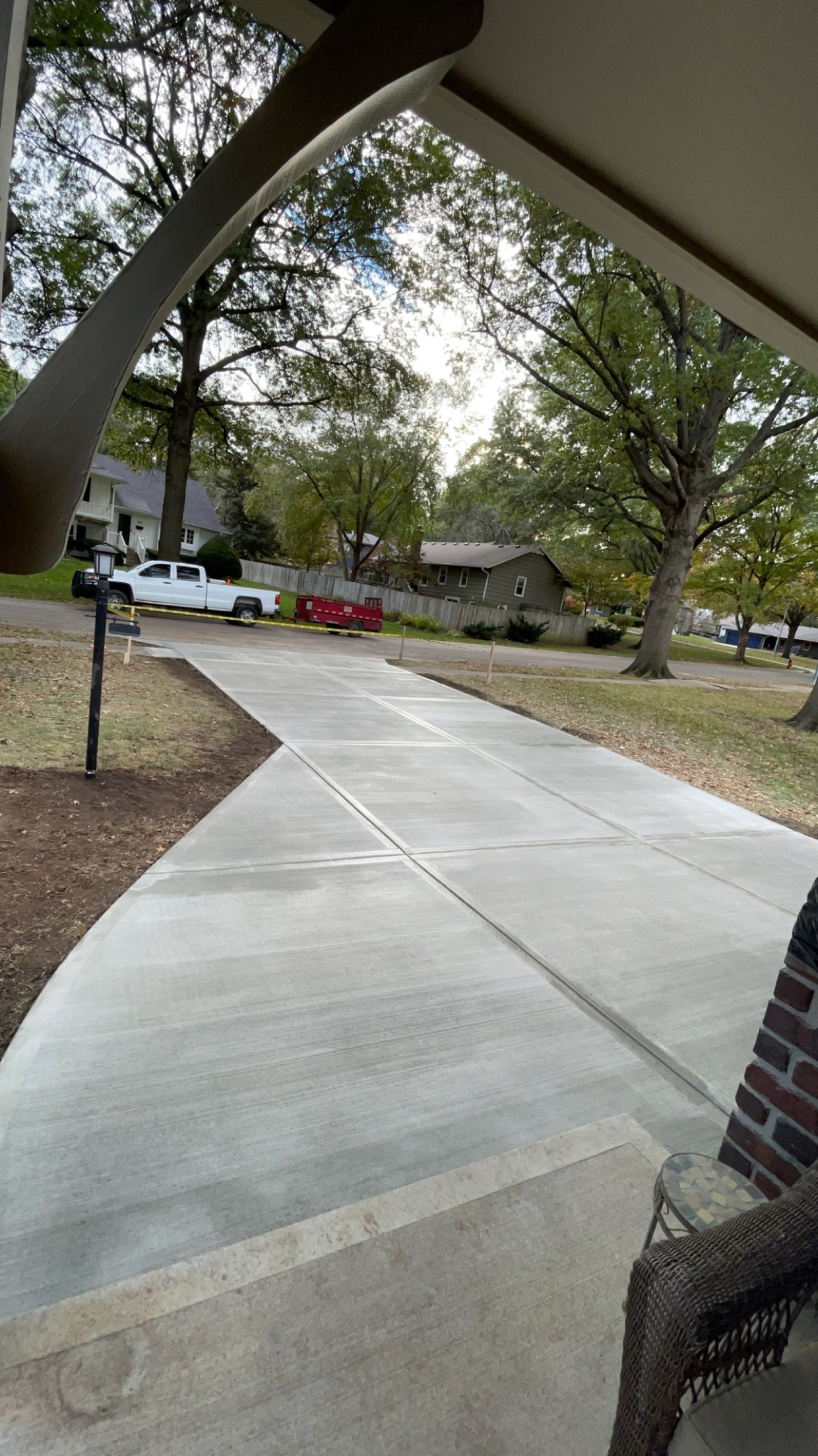 View from a covered porch down a finished concrete walkway toward the street, framed by autumn trees.